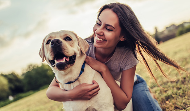 Woman with long hair crouches down to hug her dog in a field