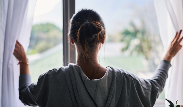 A woman seen from behind stands before a window and opens the curtains