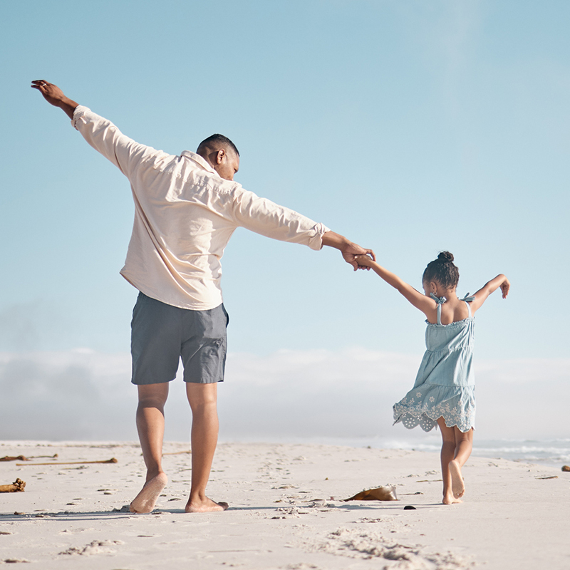 Father and young daughter hold hands as they play on a beach under a blue sky